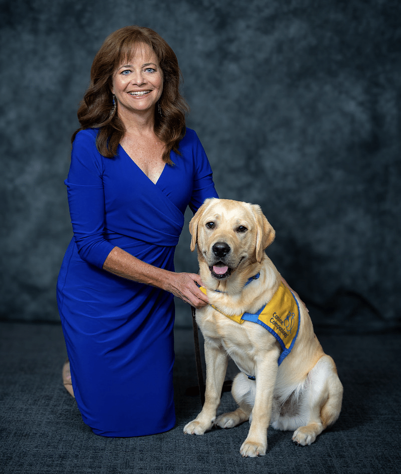 A woman wearing a blue dress kneeling next to a yellow Labrador retriever wearing a yellow and blue vest against a dark grey background. Andrea McCarren & puppy Pilot.